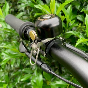 May include: A black bicycle bell with a gold-colored mounting bracket and a silver-colored clip. The bell is attached to a black bicycle handlebar. The bell has the words "Love you" engraved on it.