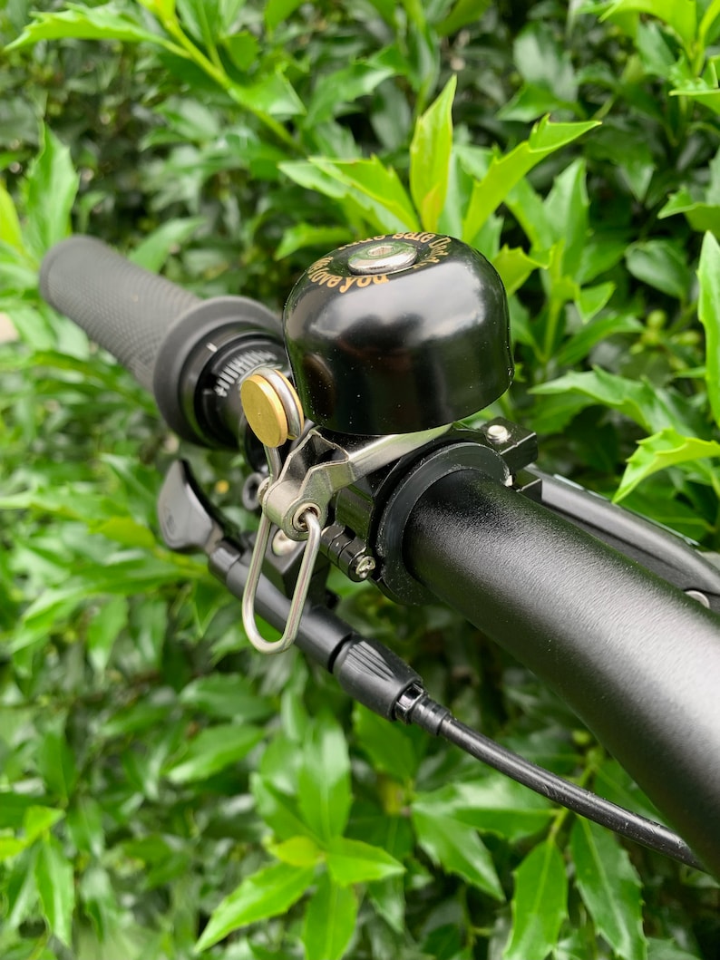 May include: A black bicycle bell with a gold-colored mounting bracket and a silver-colored clip. The bell is attached to a black bicycle handlebar. The bell has the words "Love you" engraved on it.