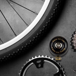May include: A black bicycle bell with gold lettering that reads "Enjoy the ride. I love you Travis". The bell is on a gray surface with a bicycle chain and a bicycle wheel in the background.
