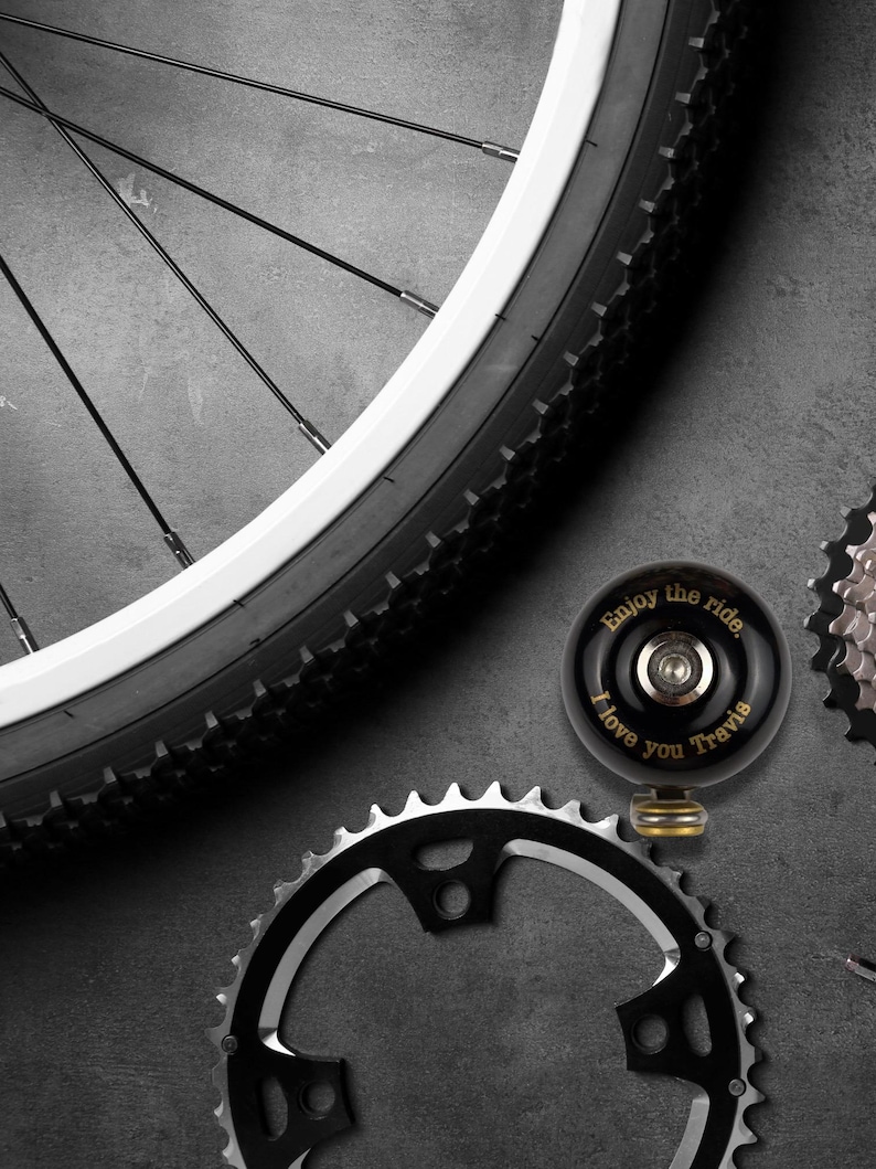 May include: A black bicycle bell with gold lettering that reads "Enjoy the ride. I love you Travis". The bell is on a gray surface with a bicycle chain and a bicycle wheel in the background.