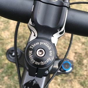 May include: Close-up of a black bicycle stem with a silver top cap. The top cap has the text "These pretzels are making me thirsty" engraved on it. The stem is attached to a black handlebar with a gray frame.