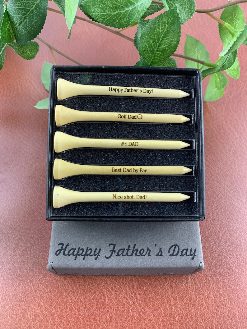 May include: A set of five wooden golf tees in a black box. The tees are engraved with messages for Father's Day, including "Happy Father's Day!" "Golf Dad" "#1 DAD" "Best Dad by Par" and "Nice shot, Dad!" The box is also engraved with the text "Happy Father's Day".