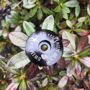 May include: Black bike stem cap with white text that reads "2.4 Swim 112 Bike 26.2 Run".