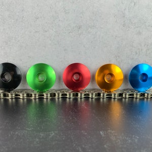 May include: Five bicycle headset top caps in different colors: black, green, red, orange, and blue. The caps are arranged in a row on a silver chain.