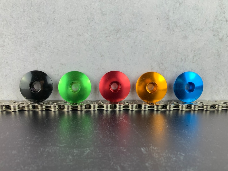May include: Five bicycle headset top caps in different colors: black, green, red, orange, and blue. The caps are arranged in a row on a silver chain.