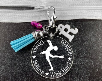 May include: A silver charm with a blue tassel, pink crystals, and a white letter "B". The charm features a silhouette of a figure ice skating with the words "Dream Believe Work Hard Achieve" around the edge.