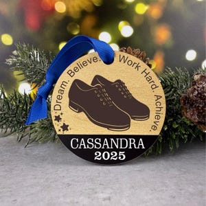 Peut inclure: Ornement de Noël rond en bois avec un ruban bleu. Il présente une paire de chaussures marron, les mots "Dream. Believe. Work Hard. Achieve." et le nom "Cassandra" avec l'année "2025".