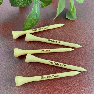 May include: Four wooden golf tees with personalized messages. The messages include "Happy Father's Day!", "Golf Dada", "#1 Dada", "Best Dada by Par", and "Nice shot, Dada!"