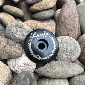 May include: Black bicycle stem cap with a silver engraved logo that reads "Leeds Cyclery". The logo includes a stylized bicycle chain ring and a horseshoe.