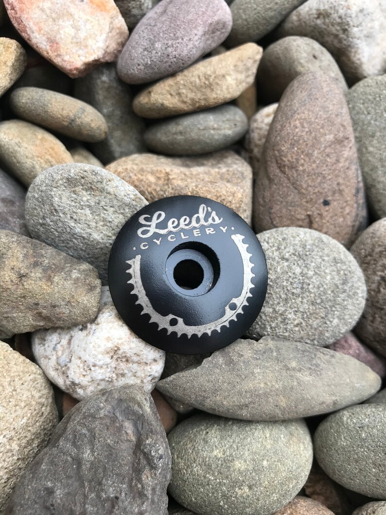 May include: Black bicycle stem cap with a silver engraved logo that reads "Leeds Cyclery". The logo includes a stylized bicycle chain ring and a horseshoe.