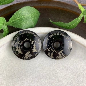 May include: Two black bicycle stem caps with engraved mountain scenes and inspirational quotes. The first cap reads "I can do all things through Christ who strengthens me". The second cap reads "Never take it out of Turbo!"