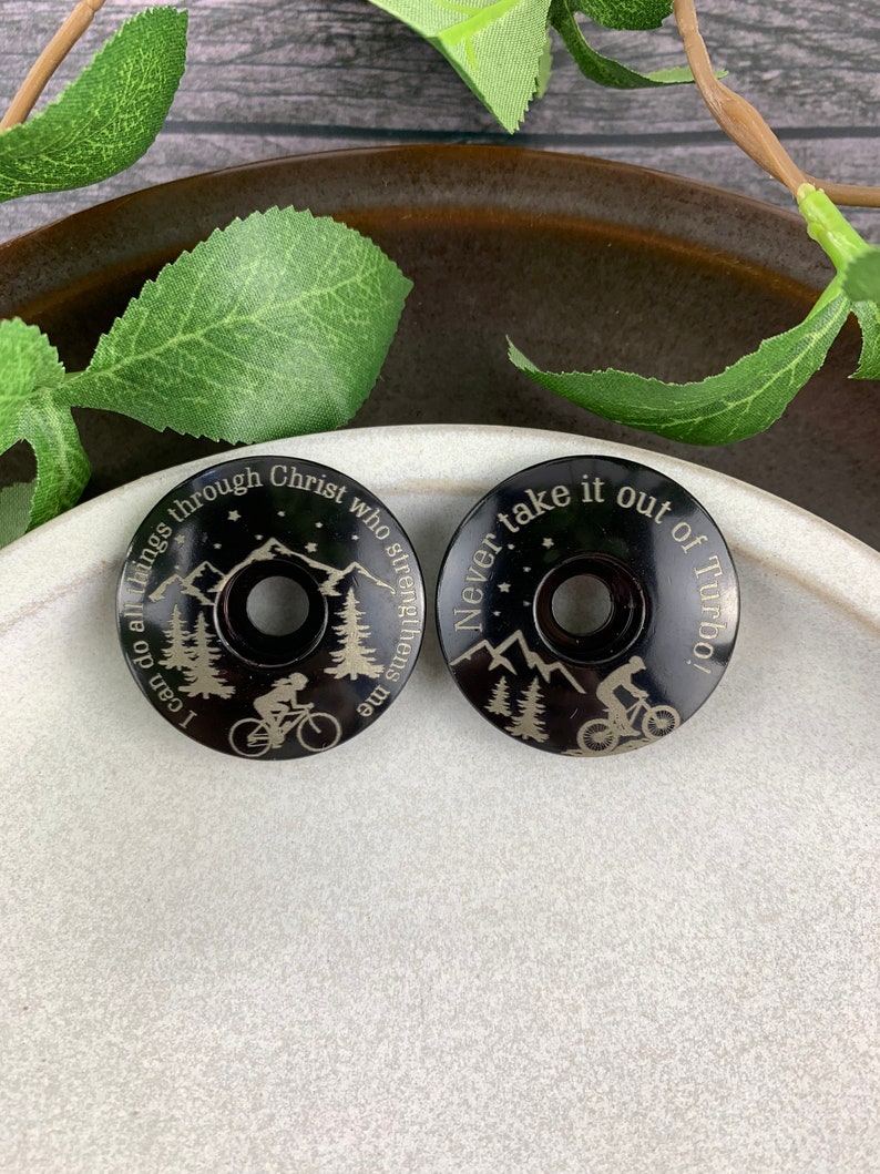 May include: Two black bicycle stem caps with engraved mountain scenes and inspirational quotes. The first cap reads "I can do all things through Christ who strengthens me". The second cap reads "Never take it out of Turbo!"