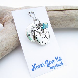 May include: Silver bag charm with a soccer ball, a star, and a blue crystal charm. The charm is attached to a silver clip. The charm has the words "Never Give Up" written on it.