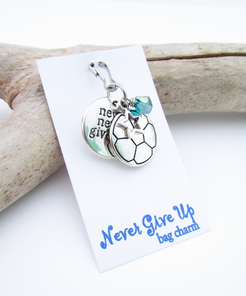 May include: Silver bag charm with a soccer ball, a star, and a blue crystal charm. The charm is attached to a silver clip. The charm has the words "Never Give Up" written on it.