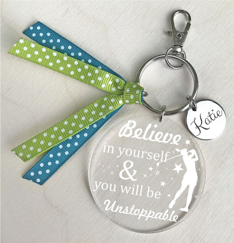 May include: A white acrylic keychain with the text "Believe in yourself & you will be Unstoppable" and a silhouette of a woman playing golf. The keychain has a green and blue polka dot ribbon attached and a silver keyring with a silver charm that says "Katie".