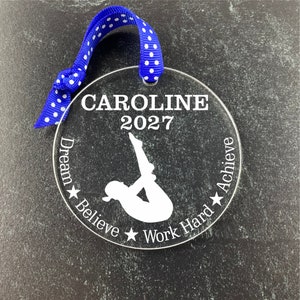 May include: A clear round ornament with a white silhouette of a gymnast performing a handstand. The ornament has the text "CAROLINE 2027" and the words "Dream Believe Work Hard Achieve" around the edge.