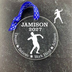 May include: A clear acrylic disc ornament with a blue and white polka dot ribbon. The ornament features the name "JAMISON" and the year "2027" in white text, along with a silhouette of a shot put athlete. The words "Dream, Believe, Work Hard, Achieve" are also inscribed.