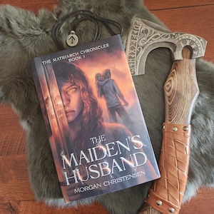 Peut inclure: Un livre intitul&eacute; "The Maiden's Husband" avec une femme sur la couverture, une hache envelopp&eacute;e de cuir et un collier pendentif sur une surface en fausse fourrure. La hache pr&eacute;sente des gravures complexes. Le livre fait partie de "The Matriarch Chronicles".