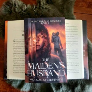 Peut inclure: Un livre intitul&eacute; "The Maiden's Husband" de Morgan Christensen, faisant partie de "The Matriarch Chronicles". La couverture pr&eacute;sente un portrait de femme et un couple en arri&egrave;re-plan. Des livres ouverts flanquent le livre principal.