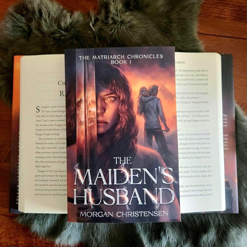 Peut inclure: Un livre intitul&eacute; "The Maiden's Husband" de Morgan Christensen, faisant partie de "The Matriarch Chronicles". La couverture pr&eacute;sente un portrait de femme et un couple en arri&egrave;re-plan. Des livres ouverts flanquent le livre principal.