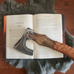 Peut inclure: Une hache argent&eacute;e orn&eacute;e d'un motif grav&eacute; est pos&eacute;e sur un livre ouvert. La hache a un manche en bois et une poign&eacute;e envelopp&eacute;e de cuir. Les pages du livre sont visibles, avec du texte et le titre du chapitre "Bai".