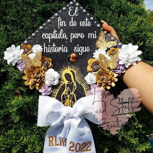 May include: Black graduation cap decorated with gold and white paper flowers, gold butterflies, and the text "El fin de este capitulo, pero mi historia sigue." A white ribbon with "RLHS 2022" is attached.