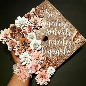 May include: A gold glitter graduation cap decorated with paper flowers in white, pink, and brown, and butterfly embellishments. The cap displays the Spanish phrase "Si puedes soñarlo puedes lograrlo."