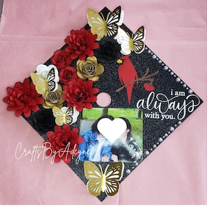 May include: A black graduation cap decorated with red, gold, and black paper flowers, gold butterflies, and a red cardinal. The cap features a photo and the text "I am always with you."