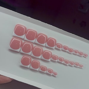 May include: A set of press-on toenails arranged in three rows on a white display card. Each square-shaped nail features a glossy nude pink base with a crisp white French tip design, creating a classic two-tone look across various sizes.
