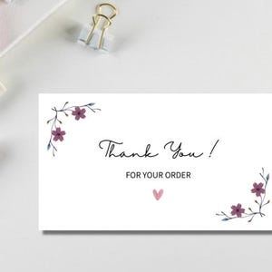 Peut inclure: Carte blanche avec l'inscription "Thank You! FOR YOUR ORDER" en écriture noire. La carte est ornée de motifs floraux dans les coins et d'un petit cœur rose. Des crayons et un trombone sont en arrière-plan.