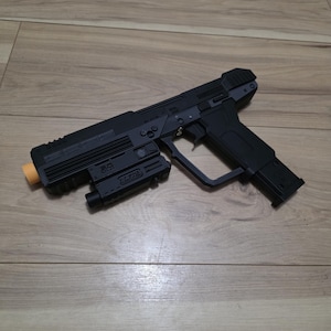 以下が含まれることがあります： マットブラックの玩具用ピストル。長方形のフレーム、トリガーガード、銃身の下に取り付けられた取り外し可能なアクセサリーが特徴です。銃口には明るいオレンジ色の先端があり、アクセサリーの側面には小さな黒い文字が記されています。