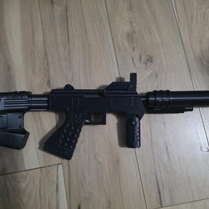 Puede incluir: Un subfusil de juguete negro que presenta una empuñadura texturizada con un pequeño emblema, una empuñadura delantera vertical, una extensión de cañón cilíndrica y una culata sólida con un patrón geométrico. El juguete tiene un acabado mate.