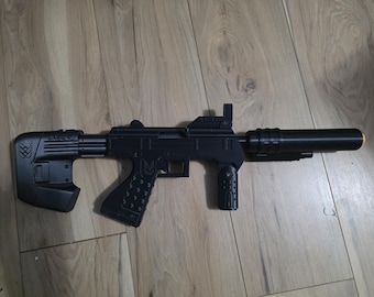 May include: A black toy submachine gun featuring a textured pistol grip with a small emblem, a vertical foregrip, a cylindrical barrel extension, and a solid stock with a geometric pattern. The toy has a matte finish and a small orange tip.