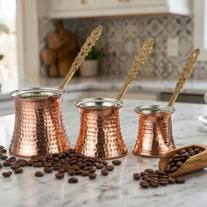 以下が含まれることがあります： 装飾的な金色のハンドルとハンマー加工されたテクスチャを備えた、3つの銅製コーヒーポット。コーヒー豆がポットの周りに散らばっており、豆の入った木製のスクープがあります。背景にはコーヒーカップがあります。