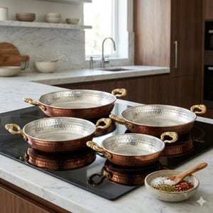 Può includere: Un set di quattro padelle color rame con interni argentati e manici dorati ornati. Le padelle sono disposte su un piano cottura nero. Una piccola ciotola di spezie e un cucchiaio di legno sono in primo piano.