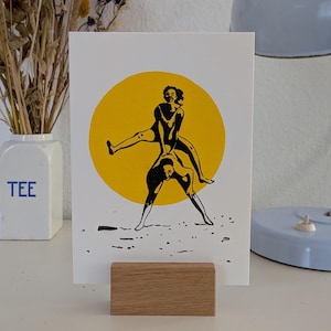 Puede incluir: Una impresión artística blanca con una ilustración en negro y amarillo de dos figuras en una pose juguetona. Las figuras están situadas contra un círculo amarillo, sujetas por un bloque de madera. Un jarrón con flores secas y una lámpara azul están en el fondo.
