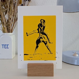 Puede incluir: Una impresión con fondo amarillo y una ilustración negra de dos figuras en una pose juguetona. La impresión se muestra en un soporte de bloque de madera. Un jarrón blanco con la palabra "TEE" es visible.