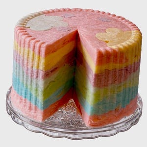May include: A colorful rainbow cake with a slice removed, revealing layers of pink, yellow, green, and blue. The cake sits on a clear glass plate and is decorated with cloud-shaped accents.