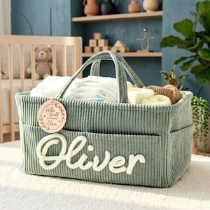 Può includere: Un contenitore per bambini in velluto a coste verde salvia con il nome "Oliver" in corsivo bianco. Il contenitore è pieno di pannolini e altri oggetti essenziali per bambini. Un'etichetta di legno recita "Hello World".