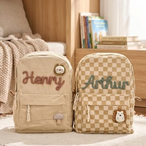 Peut inclure: Deux sacs à dos beiges. L'un est en velours côtelé avec le nom "Henry" en écriture marron et un écusson lion. L'autre est à carreaux avec le nom "Arthur" en écriture verte et un écusson hérisson. Les deux ont des poches avant.
