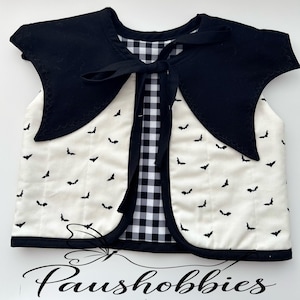 May include: A quilted vest featuring a cream-colored fabric with a black bat pattern, a solid black pointed collar with scalloped edges, and a black and white gingham lining. The garment includes a black fabric tie closure at the neckline.