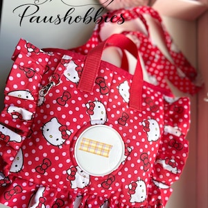 May include: A red fabric bag featuring a white polka dot pattern and Hello Kitty character graphics. The bag includes ruffled side edges, two red fabric handles, a silver metal clip, and a circular white plastic center panel with a plaid tape strip.