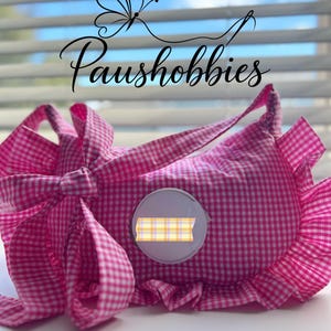 Puede incluir: Un estuche de tela con estampado de cuadros vichy en rosa y blanco, con un borde de volantes, un lazo decorativo y un parche circular blanco en el centro con una pequeña pegatina rectangular de cuadros amarillos y blancos. Texto Paushobbies.