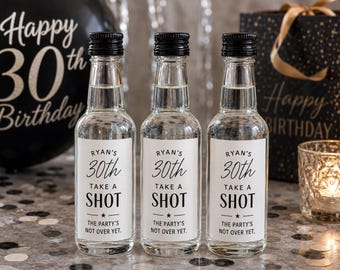 May include: Three clear glass miniature bottles with black screw caps, each featuring a white rectangular label printed with the text: RYAN'S 30th TAKE A SHOT THE PARTY'S NOT OVER YET. The bottles are filled with a clear liquid and stand upright.