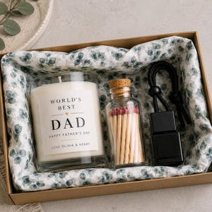 May include: A gift box containing a white candle in a glass jar with a label reading World's Best Dad, Happy Father's Day, Love Olivia & Henry, a small glass bottle filled with wooden matches, and a black car air freshener with a hanging cord.