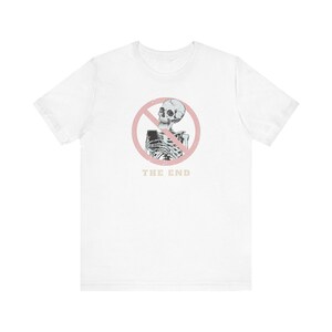 May include: White t-shirt with a graphic of a skeleton inside a pink circle with a red line through it. The text "THE END" is printed below the graphic in a light tan colour. The shirt is made of a soft material.