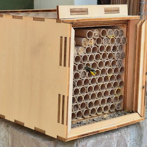 May include: A wooden bee house with a rectangular frame and a hinged door. The interior is filled with numerous cylindrical tubes, providing nesting spaces. A small bird with yellow and black markings is perched inside the house. The house is made of light-colored wood.