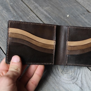 May include: Open, brown leather wallet with a bi-fold design. The interior features a series of curved, tan and brown stripes. The wallet is held open, showcasing the interior design and stitching details. A small emblem is visible.