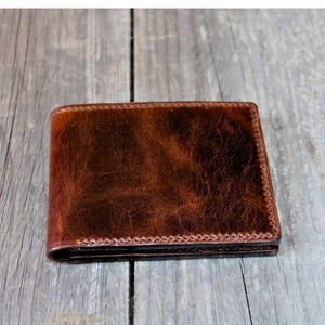 May include: A brown leather wallet with a textured surface. The wallet is rectangular and has a stitched border. The wallet is sitting on a wooden surface.
