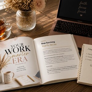 Op de afbeelding: Een open boek getiteld "Your Work Smarter Era" met een koffiemok, pen en notitieboek op een houten bureau. Een laptop met de woorden "faith focus finish" staat op de achtergrond. Een kopje thee en een vaas met bloemen zijn ook aanwezig.